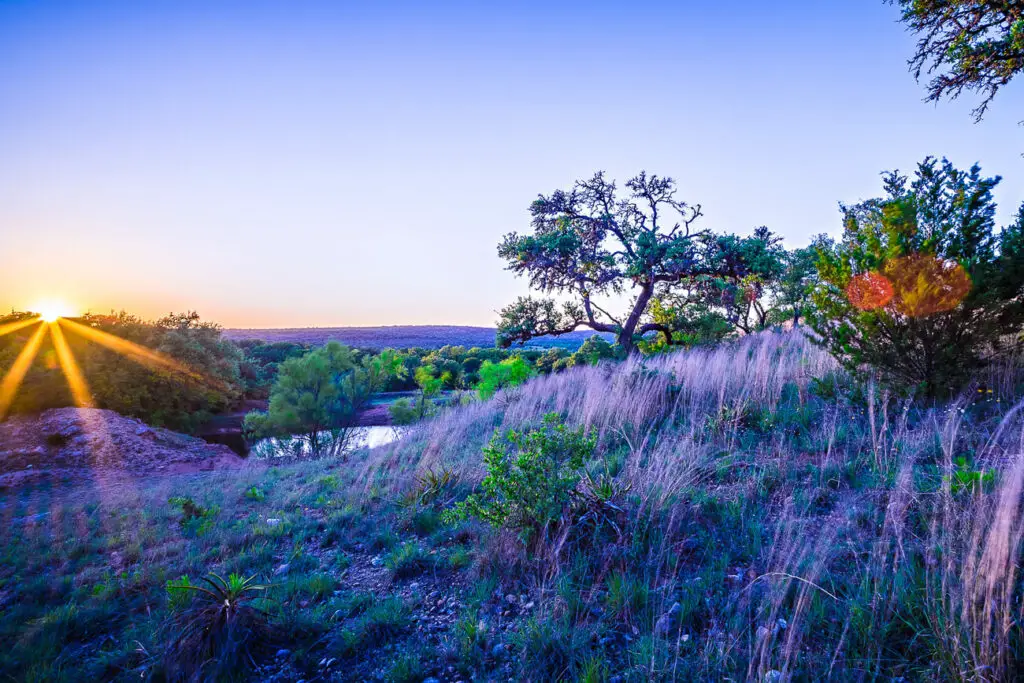 Sunset view over a tranquil Texas Hill Country landscape near Fredericksburg, with the sun casting long rays over rolling hills, a serene water body, and a variety of vegetation including tall grasses and scattered trees. One of the best things to do in Fredericksburg TX is drive the Willow City Loop.
