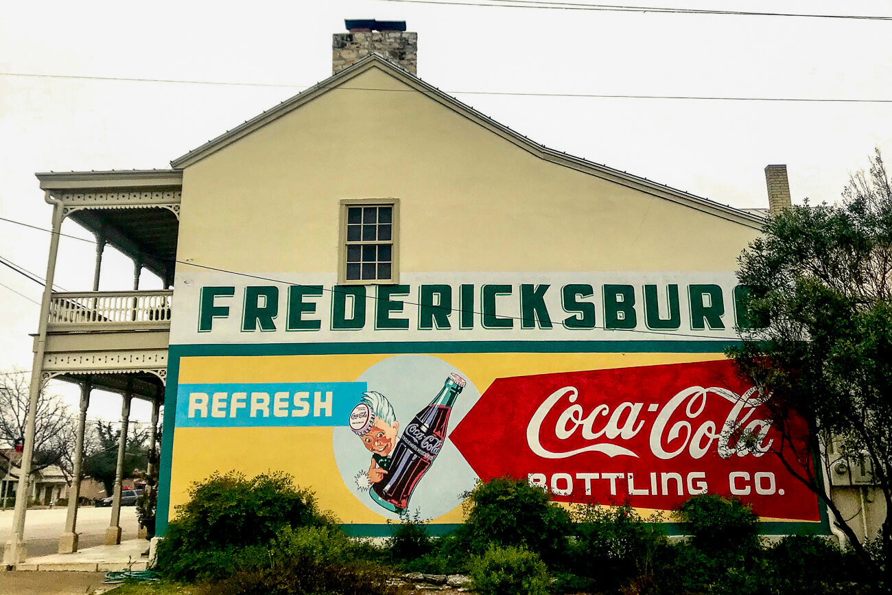 A large mural painted on the side of a building depicting a vintage Coca-Cola advertisement with 'Fredericksburg Bottling Co.' text, showcasing the town's retro Americana vibe.