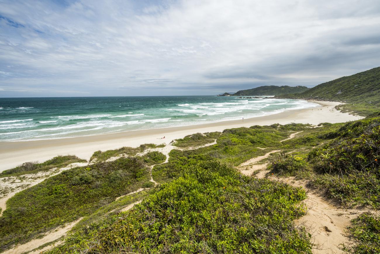 A beautiful, expansive beach scene. The foreground shows a winding sandy path leading through lush greenery and shrubs towards a wide sandy beach. The beach extends along the coast, bordered by a vibrant blue ocean with waves gently crashing onto the shore. The background features rolling hills with sparse vegetation under a partly cloudy sky.