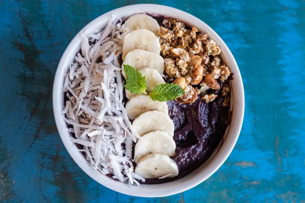 A bowl of açaí topped with sliced bananas, granola, shredded coconut, and a sprig of fresh mint, presented on a vibrant blue table surface. Acai bowls are everywhere in Ilha Grande, Brazil, and a great traditional Brazillian dish to try when you visit. 