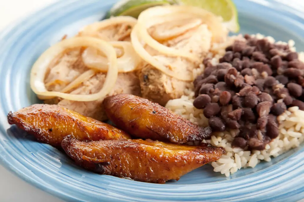 A plate featuring a serving of Cuban roast pork, topped with sautéed onions, accompanied by white rice, black beans, and sweet fried plantains.