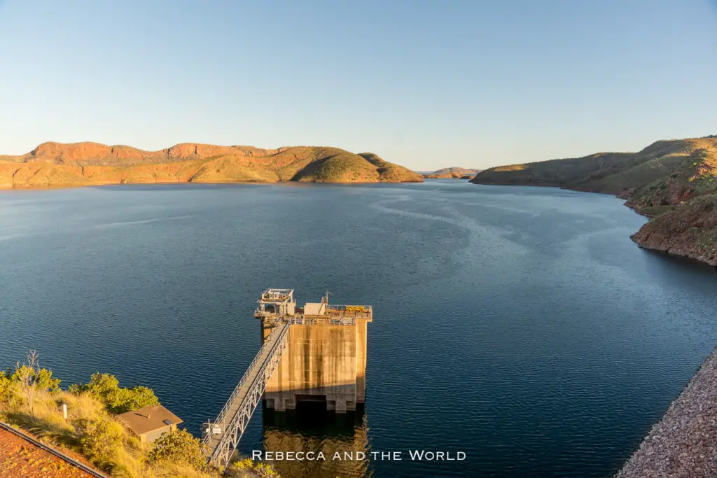 A view of the Lake Argyle Dam in Western Australia, with a large expanse of blue water surrounded by rolling hills under a clear sky. The dam structure extends into the lake, showcasing the scale and engineering of this significant site on a Darwin to Broome road trip.