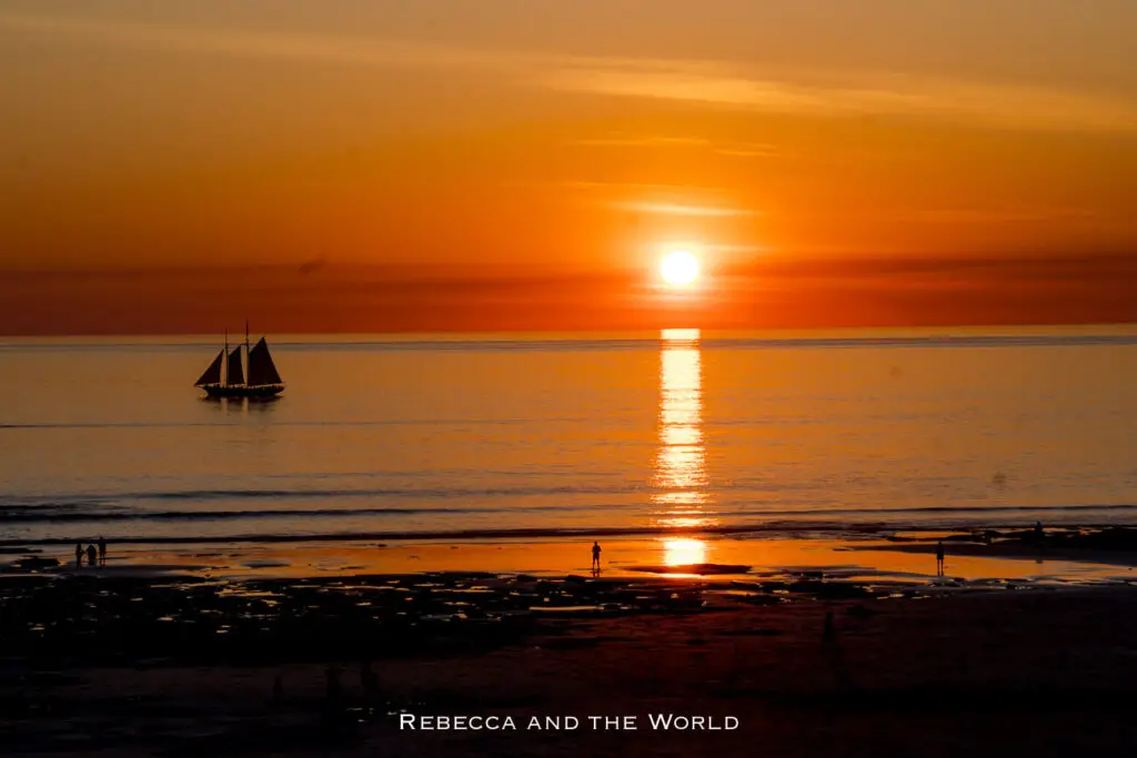A breathtaking sunset at Cable Beach, Broome, with a sailboat silhouetted against the orange and golden sky. The sun's reflection creates a shimmering path on the calm ocean, while people walk along the beach, enjoying the serene moment.
