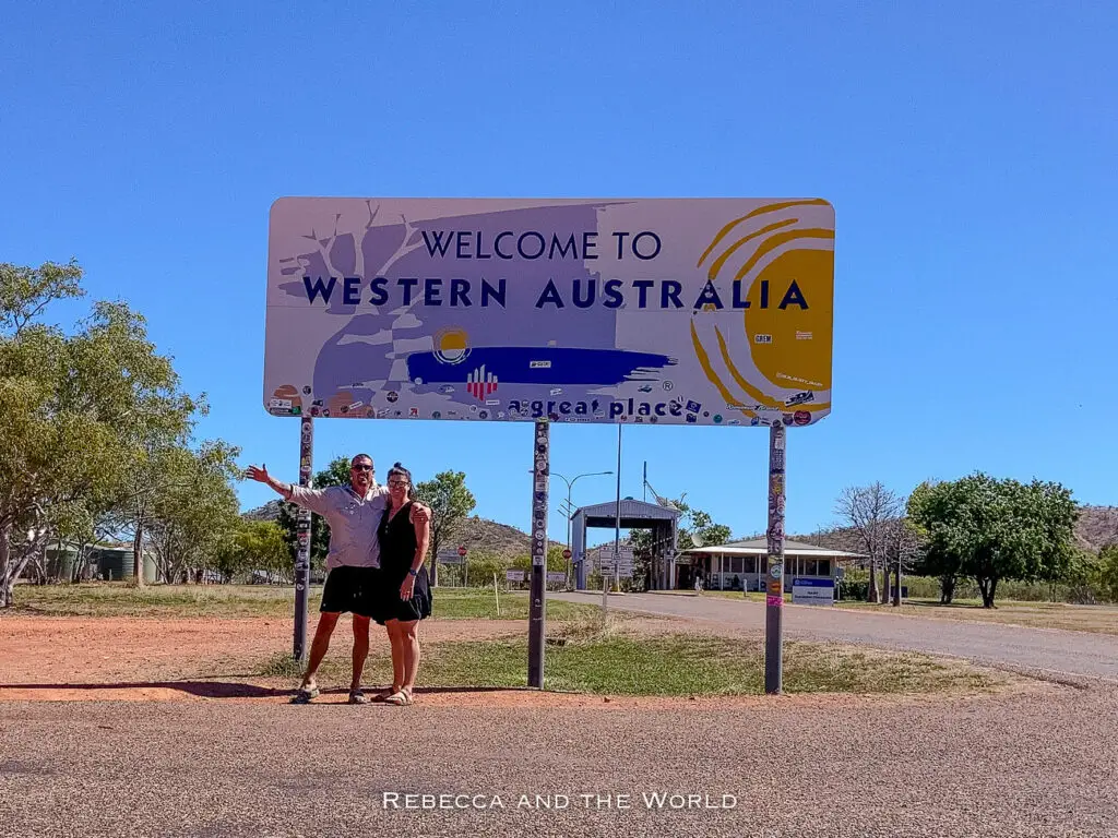 Two people - the author of this article and her husband - posing next to a large "Welcome to Western Australia" sign at the border checkpoint. The sign is colourful with blue and yellow designs, welcoming travellers embarking on a road trip from Darwin to Broome.