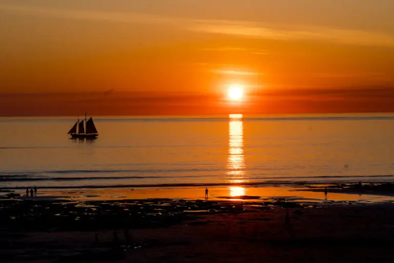A breathtaking sunset at Cable Beach, Broome, with a sailboat silhouetted against the orange and golden sky. The sun's reflection creates a shimmering path on the calm ocean, while people walk along the beach, enjoying the serene moment.