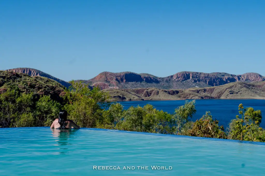 A person with long dark hair is relaxing in an infinity pool overlooking a scenic view of red rocky hills and a blue lake under a clear blue sky. This is Lake Argyle in Western Australia.