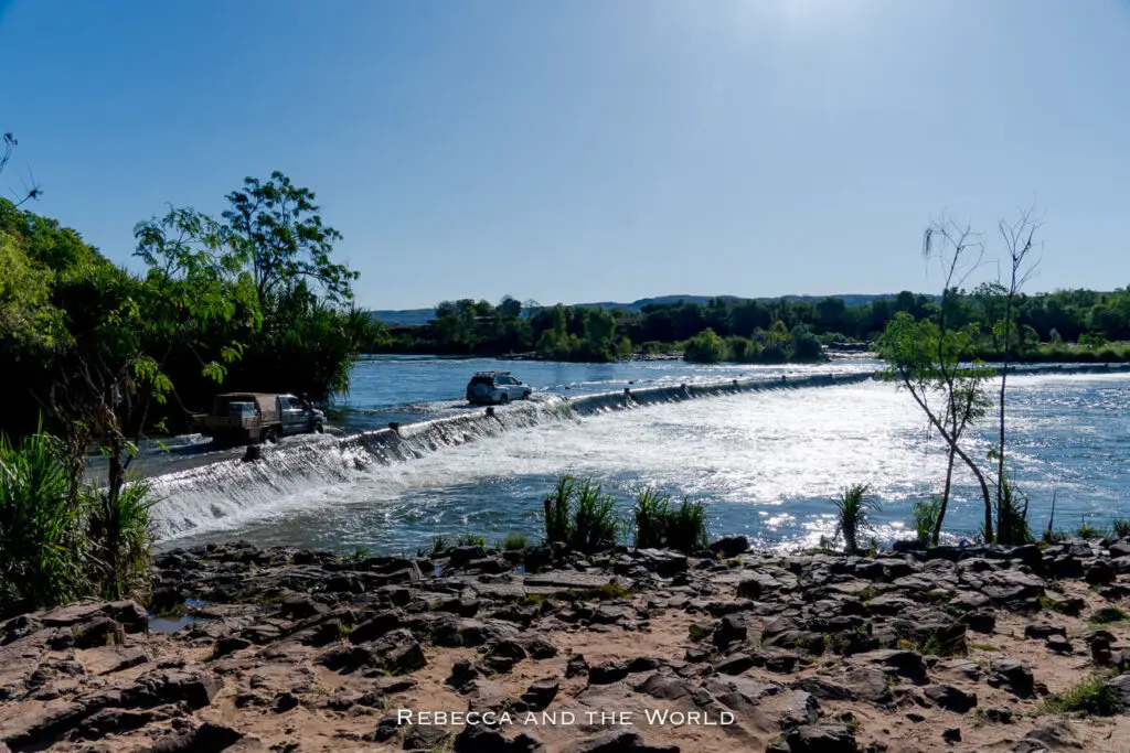 Two vehicles are driving across the causeway that crosses the Ord River, with water cascading over a low concrete causeway. Trees and vegetation line the riverbanks, and the sky is clear and bright. This is the famous Ivanhoe Crossing, one of the most popular places to see in Kununurra WA.