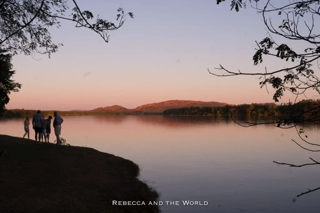 A group of people standing by Lake Kununurra at sunset. The sky is tinged with pink and orange, reflecting on the water, with tree branches framing the top of the image and hills in the background.