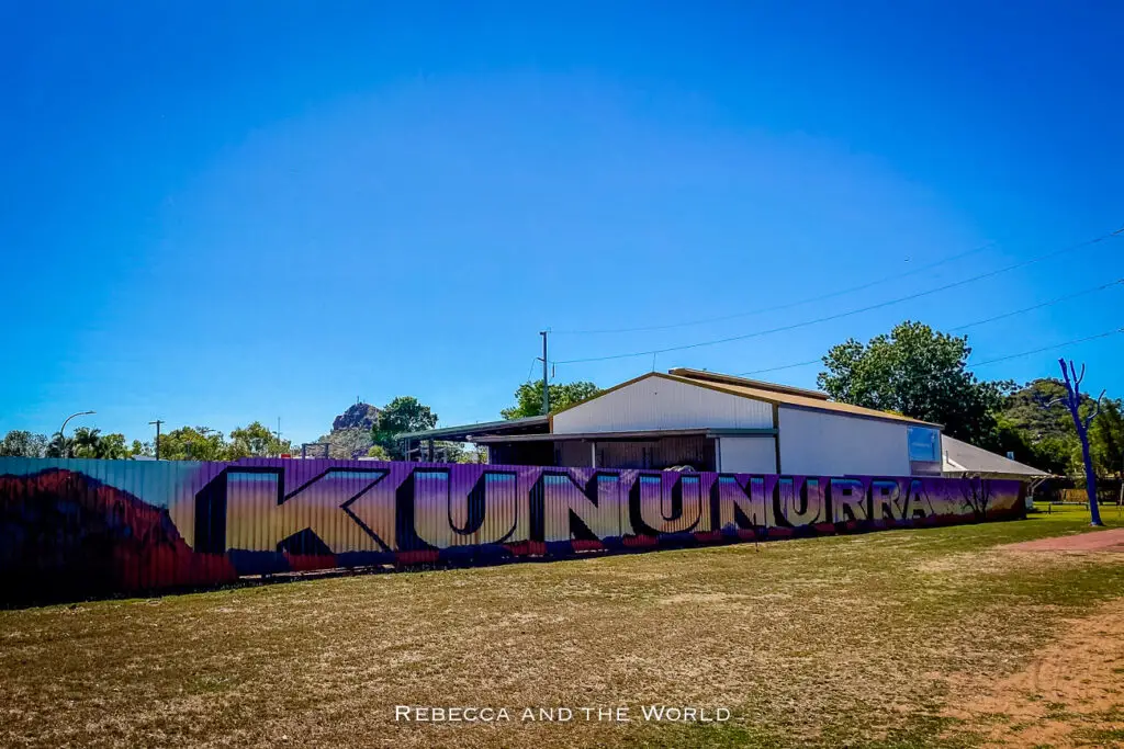 A fence with a large mural of "Kununurra" painted on the side. The mural is colourful, set against a backdrop of a clear blue sky and a grassy area in the foreground.