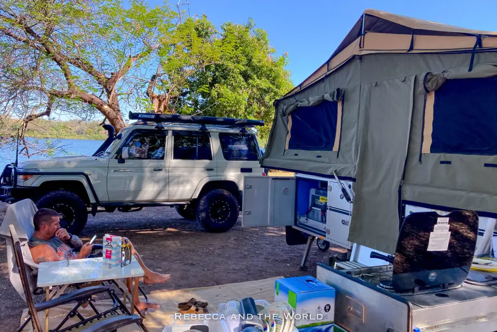 A camping setup with a man sitting in a chair, using his phone. A large off-road vehicle and a rooftop tent are parked nearby under the shade of trees by a river. This is the author's husband relaxing at the Discovery Parks Lake Kununurra.