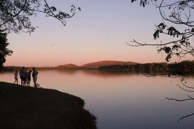 A group of people standing by Lake Kununurra at sunset. The sky is tinged with pink and orange, reflecting on the water, with tree branches framing the top of the image and hills in the background.