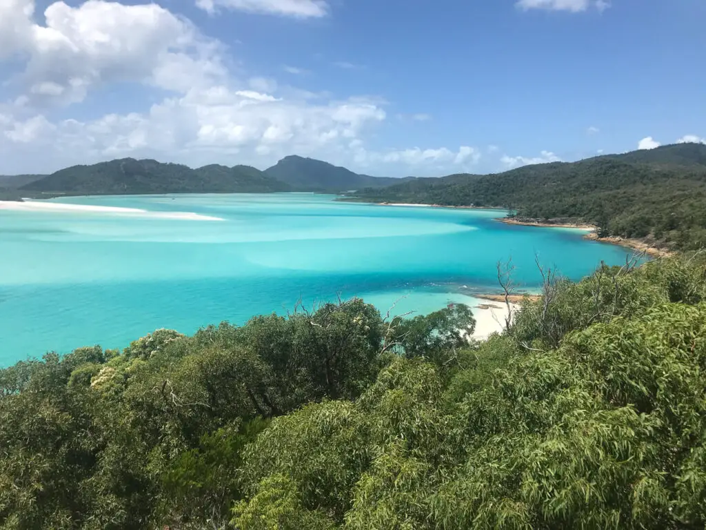 A stunning view of turquoise blue waters with white sandbars and green islands in the background, taken from an elevated vantage point on the Whitsunday Islands.