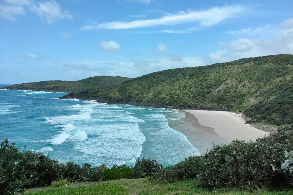 A scenic coastal view of waves crashing onto a sandy beach surrounded by lush green hills, under a blue sky with some clouds. One of the best Australian road trips is up the East Coast of Australia from Melbourne to the Gold Coast.