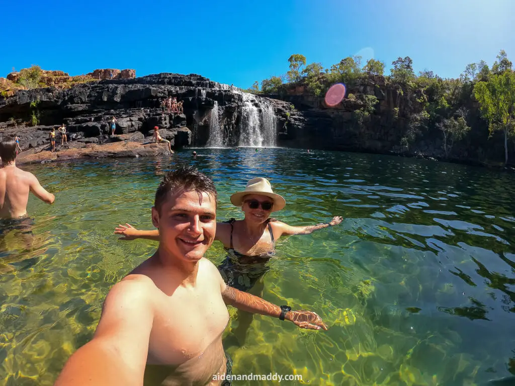 A group of people swimming in the clear, green waters of a natural pool at Manning Gorge on the Gibb River Road, with a rocky cliff and cascading waterfall in the background under a bright blue sky.