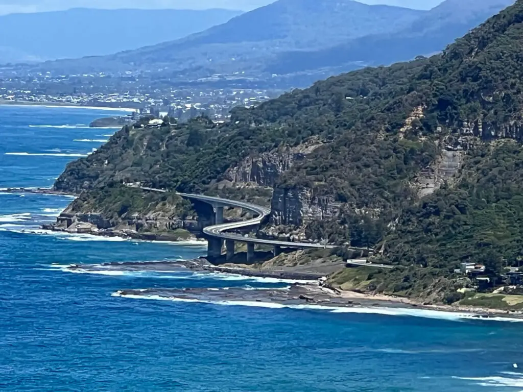 A curving coastal bridge (Sea Cliff Bridge) built along a steep, forested cliff overlooking the ocean, with mountains in the distance and a coastal town visible in the background. The Grand Pacific Drive is one of the best Australian road trips, easily drivable in a day.