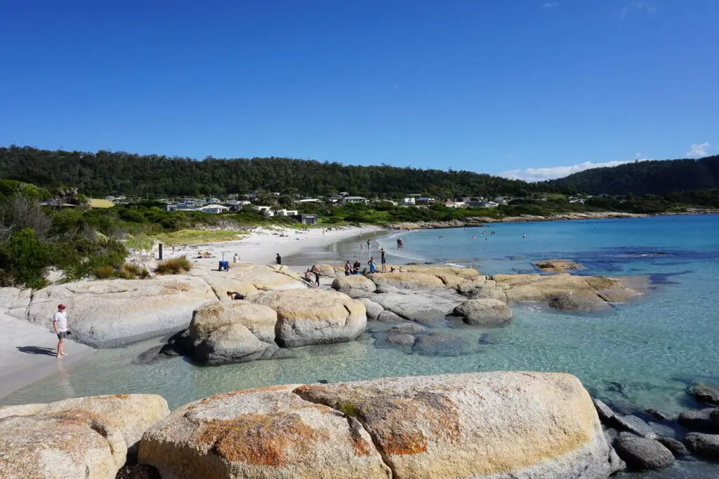 A beach scene at the Bay of Fires featuring large granite boulders with orange lichen, clear shallow water, and people enjoying the sandy shoreline with green hills and a few houses in the background. A road trip along the East Coast of Tasmania is a great option, as the island state is so compact and easy to drive.