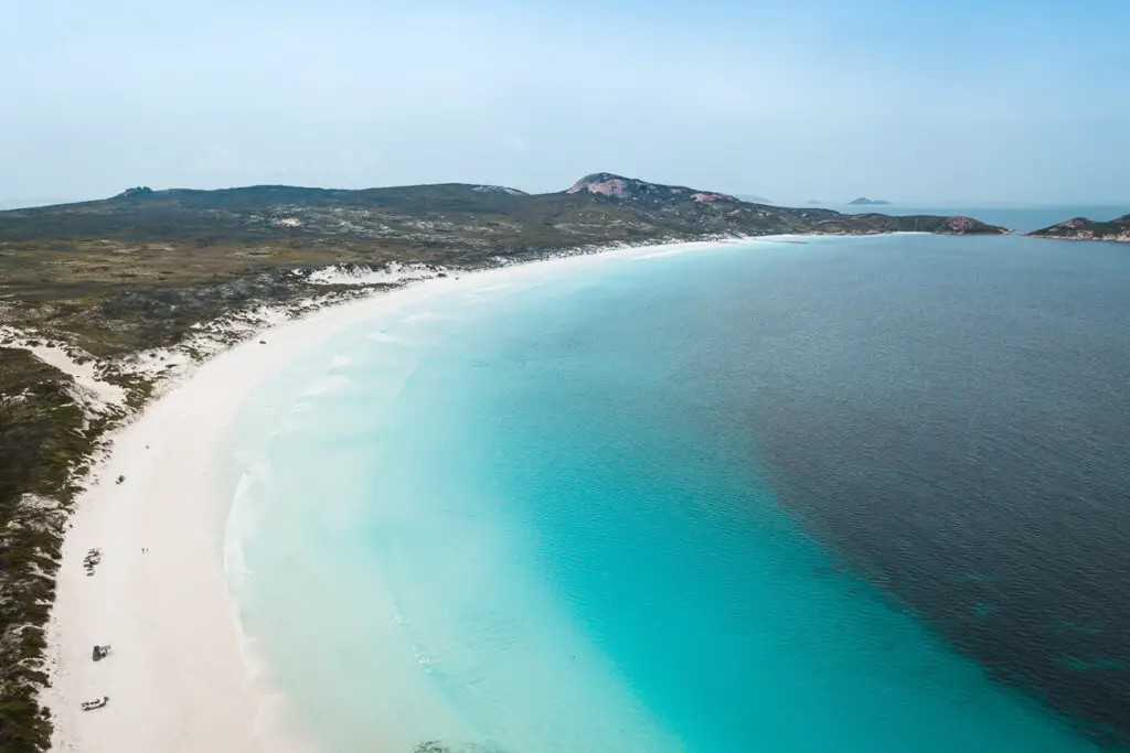 A panoramic view of a pristine white-sand beach with gentle turquoise waves, stretching along the shoreline with sparse vegetation and low hills in the background. Lucky Bay in Western Australia is one of the most beautiful stops on a Perth to Esperance road trip.