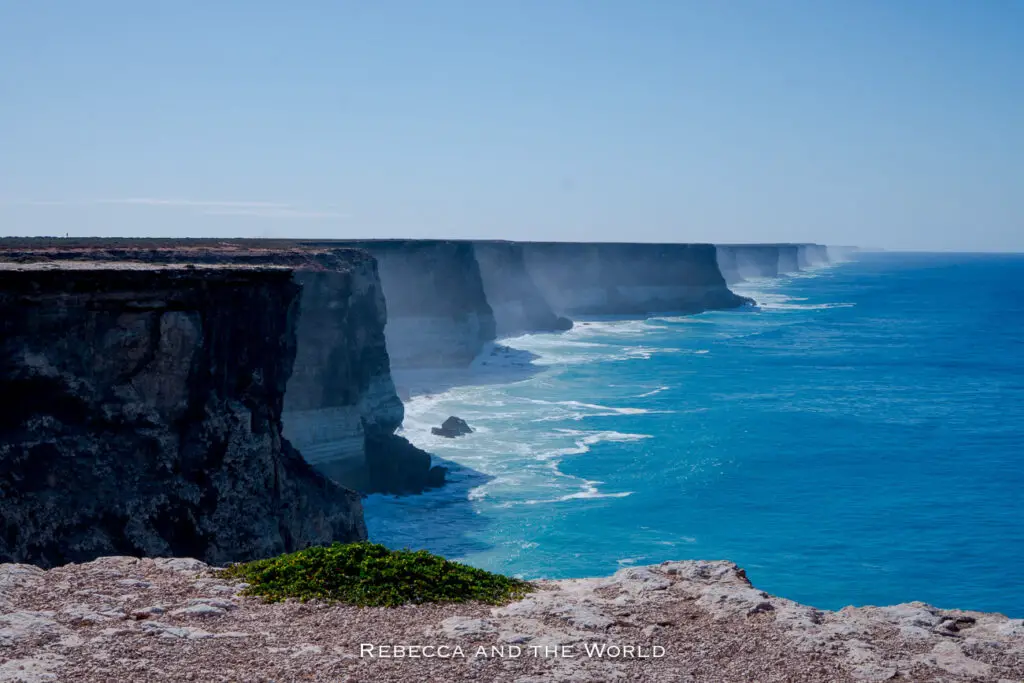 The image shows the dramatic coastline along the Great Australian Bight, with tall, steep cliffs dropping down to the blue ocean. The cliffs stretch far into the distance, with mist rising where the waves crash against the rocks. The water is a vibrant blue, contrasting sharply with the dark cliffs, creating a striking natural scene. Stop when crossing the Nullarbor at one of the lookouts.