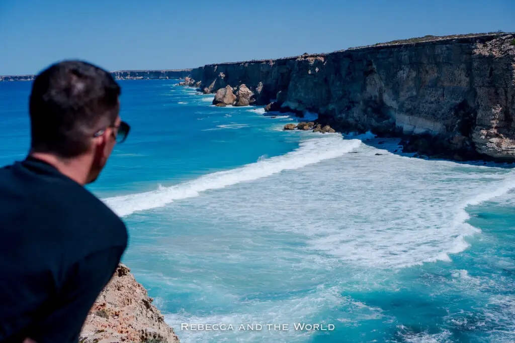 In this image, a person is seen from behind, leaning on a rocky edge as they look out over a stunning coastline. The cliffs rise steeply from the turquoise ocean, with waves crashing against the base. The coastal cliffs extend into the distance, creating a rugged and scenic view. The person is wearing sunglasses and is focused on the natural beauty ahead.