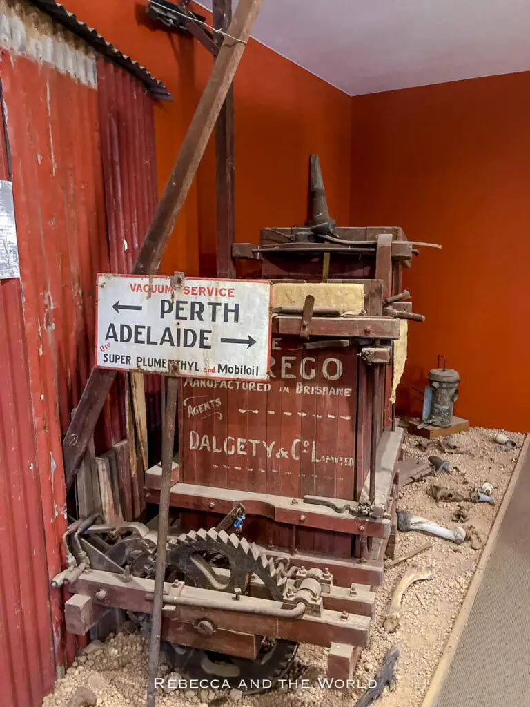 This image shows an old-fashioned piece of equipment or machinery on display in a museum. The wooden structure, surrounded by gravel and various old tools, includes a vintage sign pointing directions to "Perth" and "Adelaide," referencing historical travel or service routes. The backdrop features corrugated metal and warm red and orange walls, adding to the rustic, historical atmosphere.