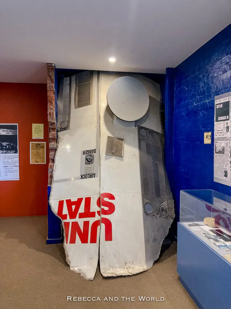 The photo features a large, metallic object mounted against a wall inside a museum or exhibit space. The object, possibly a part of a spacecraft or large vehicle, is marked with the words "UNITED STATES" upside down, with the word "DANGER" also visible. The surrounding walls are painted in bold blue and red colours, and display boards with information are partially visible on the walls. Stop at the small museum inside the Balladonia Roadhouse when crossing the Nullarbor.