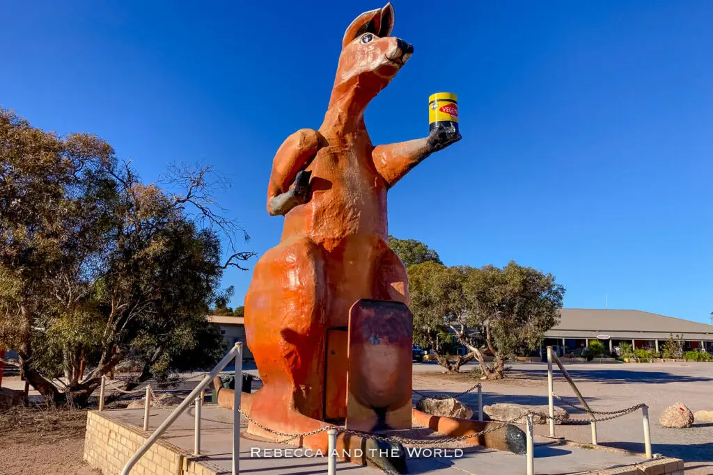 The image showcases a giant statue of a kangaroo holding a jar of Vegemite, a popular Australian spread. The statue is located outdoors, surrounded by trees and a clear blue sky, with a small building partially visible in the background. The oversized kangaroo is painted in earthy tones, adding a whimsical and distinctly Australian character to the scene. This kangaroo can be seen at Border Village, a town people pass through when crossing the Nullarbor.