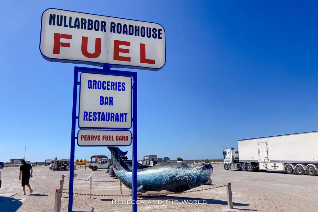 The image captures the Nullarbor Roadhouse, a fuel station and rest stop in a remote area. The large sign advertises "Fuel," "Groceries," "Bar," and "Restaurant," with a model of a whale on display below the sign. The scene includes a few parked trucks and a clear blue sky.