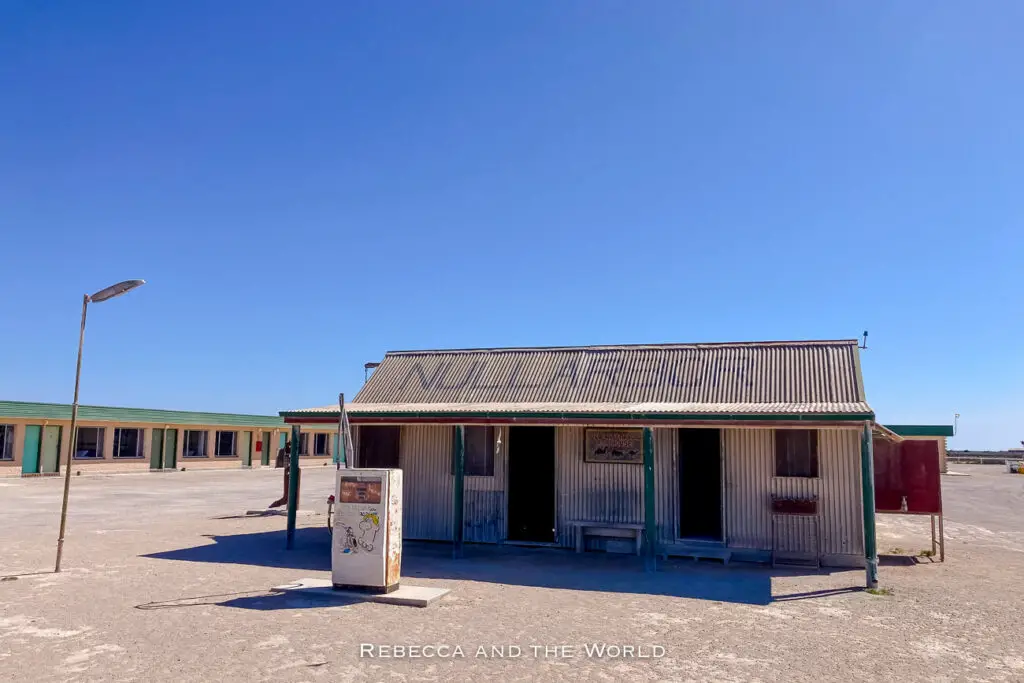 This photo shows a small, weathered building with corrugated metal walls, labelled "Nullarbor Roadhouse" in faded letters on the roof. The structure stands alone under a clear blue sky, with a dirt lot in front and a simple gas pump visible. The surrounding area is flat and barren, highlighting the remote and rugged setting of this outback roadhouse.