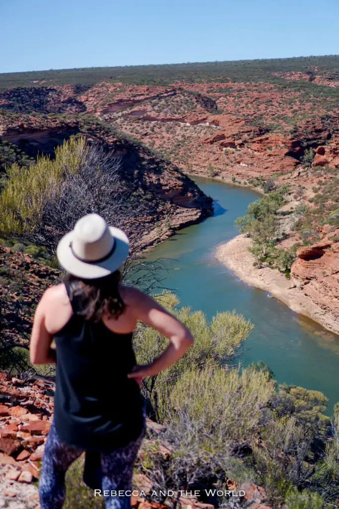 The image shows a woman - the author of this article - standing on a rocky cliff overlooking the Murchison River in Kalbarri National Park, Western Australia. She is wearing a grey brimmed hat and dark clothing, gazing out at the winding river far below. The scene captures the vast, rugged beauty of the park, with its red rock formations and deep gorges.
