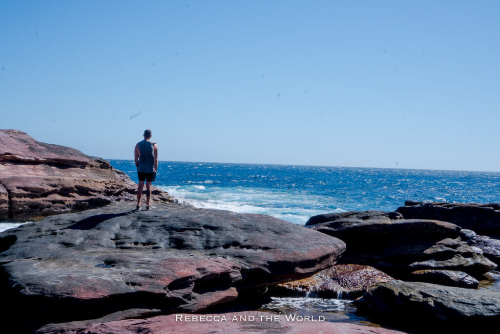 The image shows a man - the author's husband - standing on large, flat rocks overlooking the Indian Ocean at Red Bluff, near Kalbarri, Western Australia. He is gazing out at the waves crashing against the rocky shoreline, with the deep blue sea stretching to the horizon. The scene highlights the rugged coastal landscape and the powerful ocean, offering a sense of solitude and connection with nature.