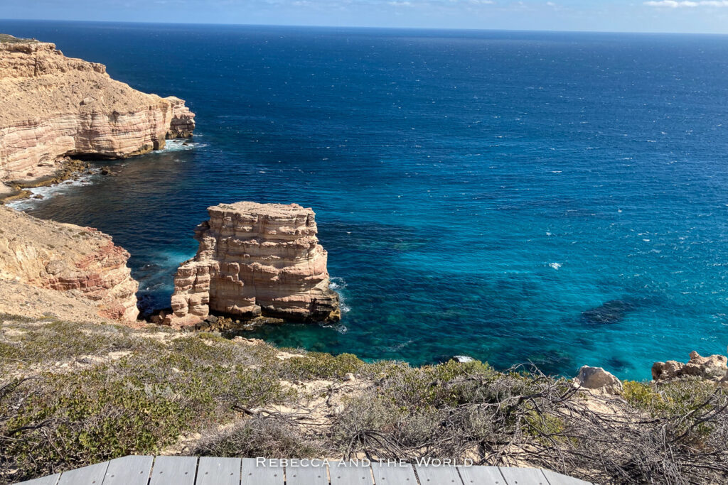 The photo captures the stunning coastal cliffs at Kalbarri National Park, Western Australia. The cliffs are made of layered beige and red rock, towering over the deep blue waters of the Indian Ocean below. The clear, vibrant water contrasts sharply with the rugged, arid landscape, showcasing the dramatic meeting of land and sea.