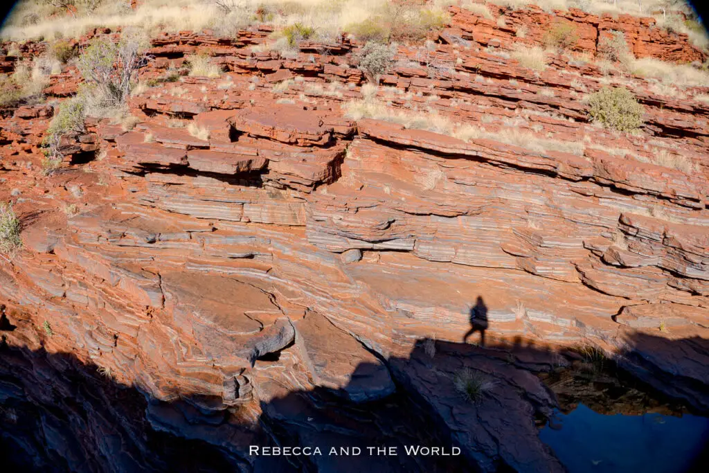 The photo highlights the rich, layered rock formations of Karijini National Park. The reddish-brown rocks are sharply defined, with visible striations that show the different layers of sediment. Sparse vegetation clings to the rock face, and a shadow of a person is cast on the rocks, giving a sense of scale to the scene.
