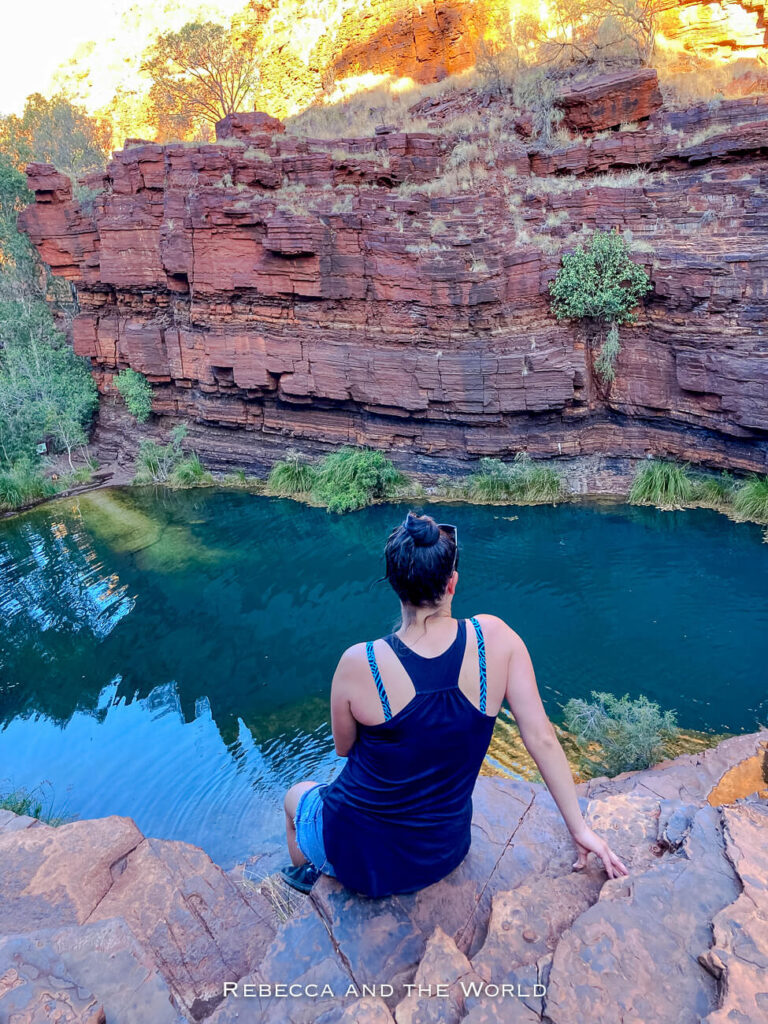 The photo features a woman sitting on the edge of a cliff overlooking the tranquil Fortescue Falls in Karijini National Park, Western Australia. The woman - the author of this article - is seated on the reddish-brown rocks, gazing out at the still, deep blue water below. Behind the waterhole, steep rock walls rise up, showcasing the park’s distinctive layered rock formations.