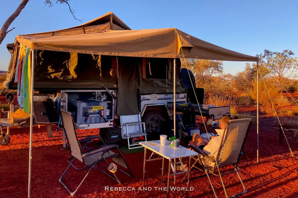 The image depicts a well-equipped camping setup in the remote, arid landscape of Karijini National Park in Western Australia. A tent attached to a trailer is set up with a canopy for shade, and camping chairs and a table are arranged outside. The warm light of the setting sun casts a golden glow over the scene, highlighting the red dirt typical of the Australian outback. The surrounding area is sparsely vegetated, with low bushes and trees scattered across the landscape.