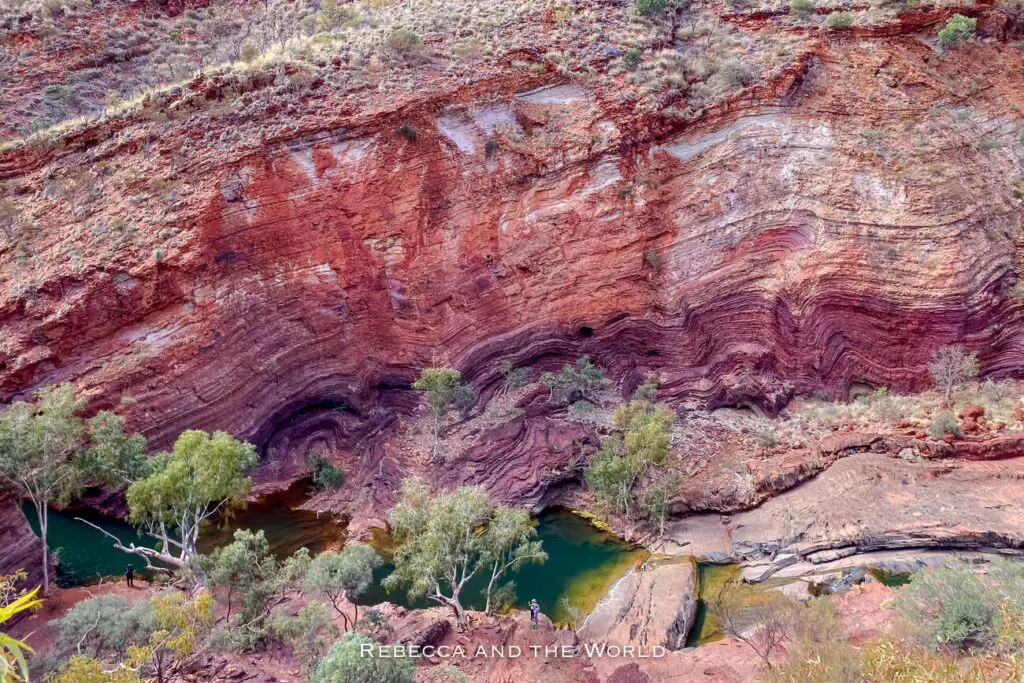 The image captures a dramatic view of Hamersley Gorge in Karijini National Park, showcasing the colourful, layered rock walls that make the park famous. The rock formations display a range of colours from deep reds to purples, with distinct lines marking the different sediment layers. At the bottom, a small pool of water is surrounded by trees and other vegetation, providing a stark contrast to the rugged, arid cliffs.