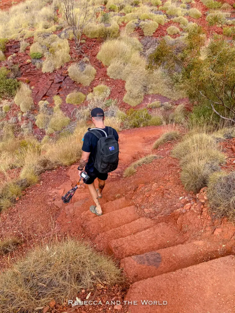 This image shows a man - the author's husband - descending a red dirt path carved into the rugged terrain of Karijini National Park. The landscape is covered with dry, grassy tufts and sparse shrubs, typical of the arid Australian outback. The man's attire suggests he is hiking; he carries a black backpack and holds a camera.
