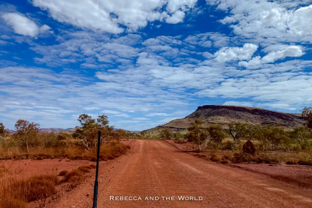 The photo captures a wide, unpaved red dirt road stretching into the distance, flanked by the arid landscape of Western Australia. The road leads toward a low mountain range, with scattered trees and shrubs lining the sides. Above, the sky is filled with white, fluffy clouds, creating a striking contrast with the deep blue sky and the red earth below.