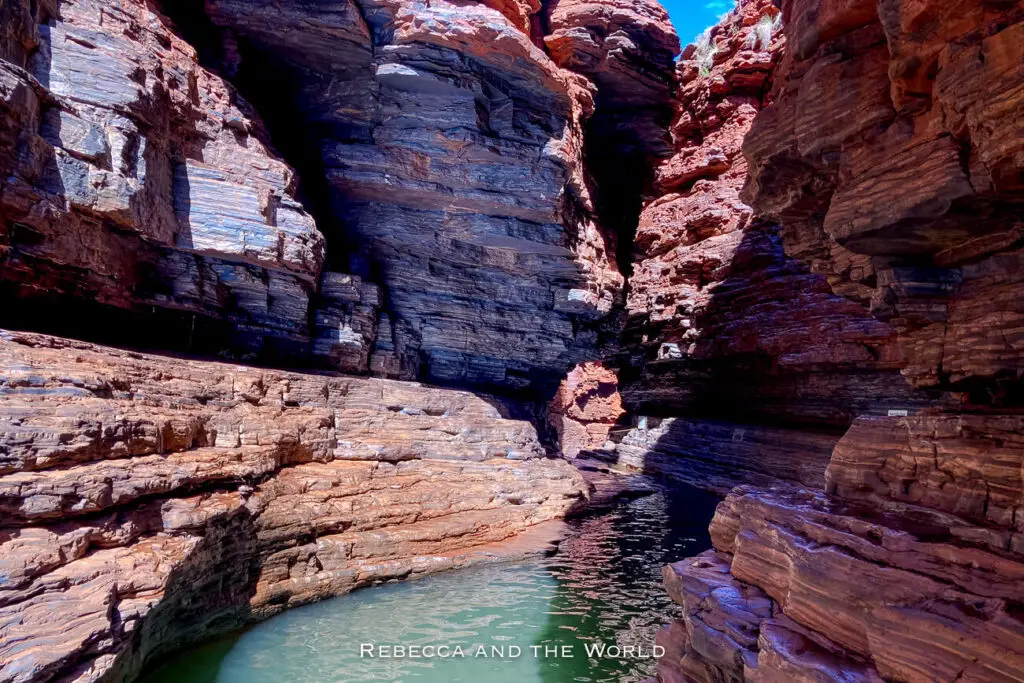 This photo depicts a deep, narrow gorge in Karijini National Park with layered rock formations on both sides. The base of the gorge is partially submerged in clear water, with light and shadow playing on the rugged rock surfaces. The dark, deep colours of the gorge contrast with the bright sunlight filtering in from above. This is Kermit's Pool.