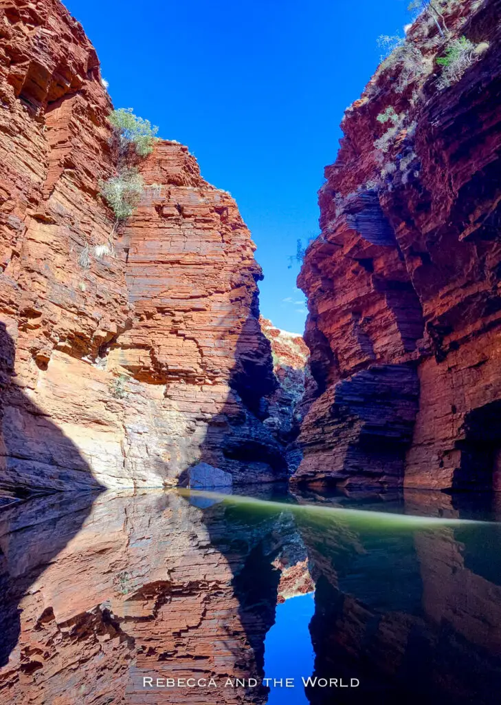 This photo shows a wide, deep gorge in Karijini National Park, with towering red rock walls that are reflected perfectly in the still water below. The blue sky above contrasts with the deep red of the rocks, creating a vivid, mirror-like effect in the water. Sparse vegetation clings to the rock walls, adding touches of green to the otherwise arid landscape. This is Handrail Pool.