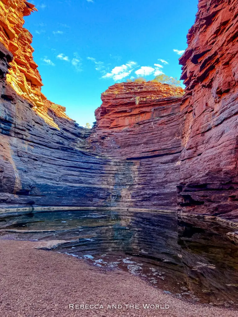 The image features a natural amphitheater-like rock formation in Karijini National Park. The curved, layered rock walls enclose a shallow pool of water, creating a serene and secluded scene. The colours range from deep red to orange and brown, with the blue sky above adding a contrasting hue. The texture and layering of the rocks highlight the geological history of the area. This is Joffre Gorge.