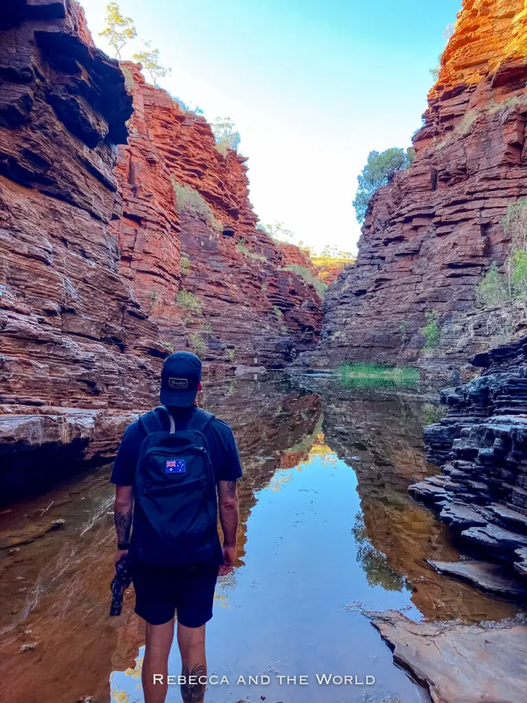 This photo shows a hiker standing at the edge of a calm pool within the narrow part of Joffre Gorge in Karijini National Park. The hiker's backpack displays an Australian flag patch, and they appear to be admiring the dramatic red rock walls that rise steeply on either side. The water reflects the vibrant colours of the rocks, creating a peaceful and contemplative atmosphere.