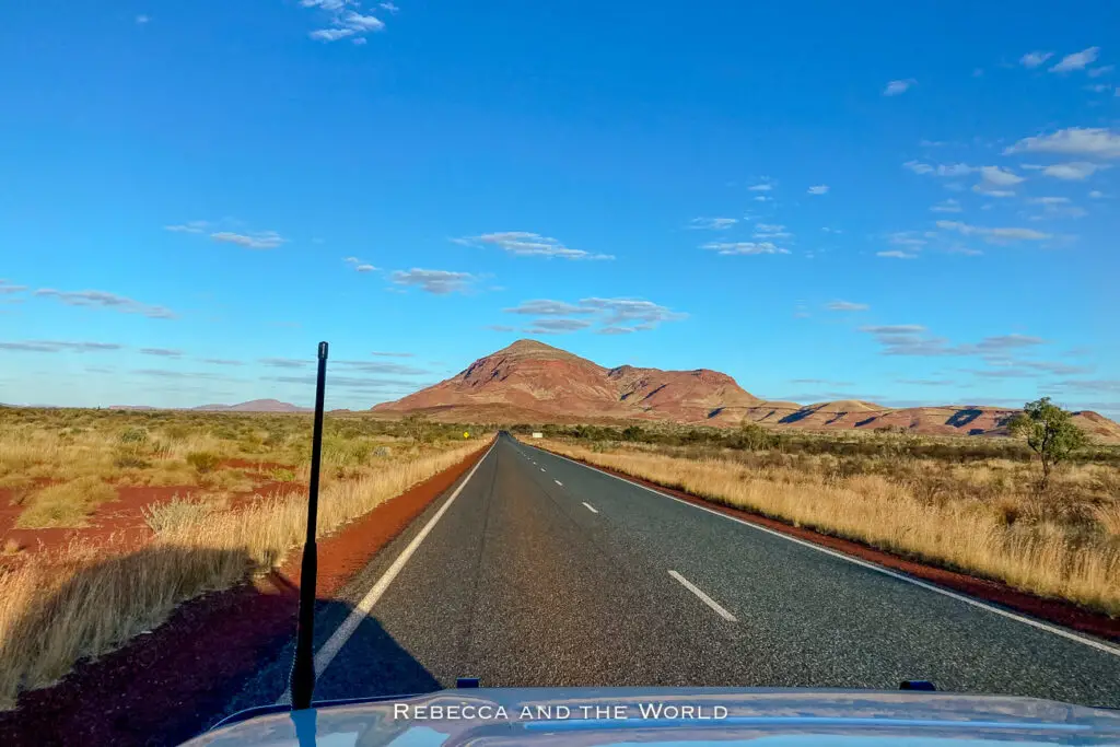 This image captures a long stretch of highway cutting through the arid landscape of Western Australia. The road leads straight toward a distant mountain, framed by wide, open plains covered in dry grasses. The sky is a brilliant blue with a few scattered clouds, and the golden light of the late afternoon casts a warm glow over the scene.