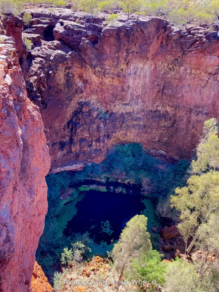 This photo offers a breathtaking aerial view of a deep, steep-walled chasm in Karijini National Park. The sheer red rock walls descend sharply to a small, dark pool at the bottom, surrounded by lush green vegetation. The contrast between the vibrant red rocks and the green plants creates a striking visual, highlighting the dramatic landscape. This is Circular Pool.