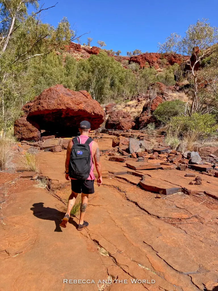 A hiker walks along a rocky trail surrounded by the red earth and green trees of Karijini National Park. The man, carrying a black backpack with an Australian flag patch, is heading toward a massive boulder that dominates the path. The surrounding landscape is rugged, with scattered trees and shrubs adding touches of green to the red-dominated scene.