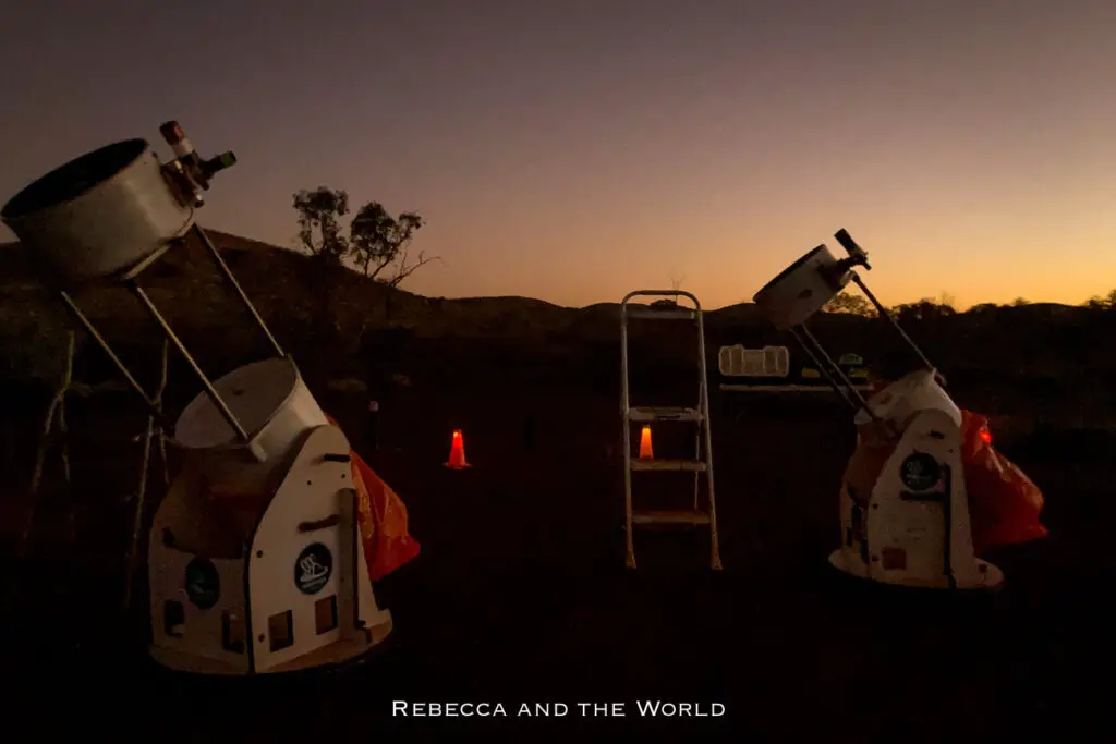 This nighttime photo shows a pair of large telescopes set up in the outback under a darkening sky, with the last light of dusk fading into the horizon. The telescopes are aimed upward, ready for stargazing, and are surrounded by faint silhouettes of trees and distant hills. The scene captures the quiet anticipation of a night spent observing the stars, with the deep twilight sky providing a perfect backdrop.