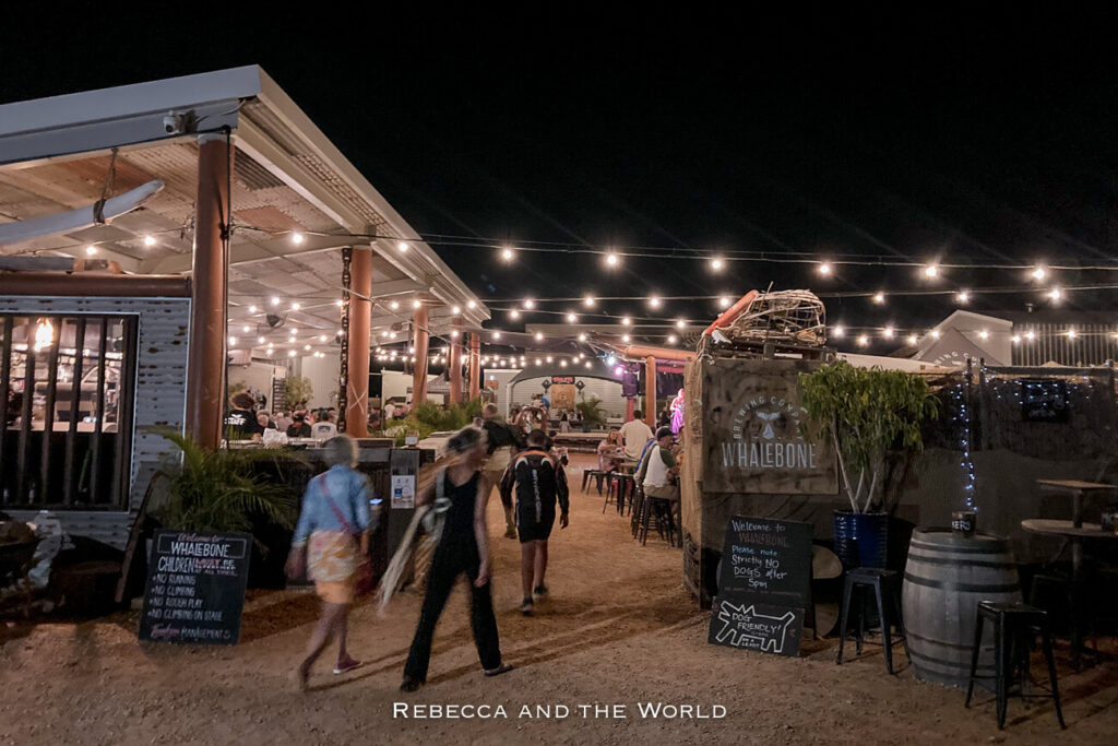 The image shows an outdoor dining area at Whalebone Brewing Company in Exmouth, Western Australia, taken at night. The scene is lit by strings of warm, glowing lights hanging overhead. Several people are seen walking towards or standing near tables where diners are seated. The setting has a casual, relaxed atmosphere with rustic decor, including barrels and potted plants. The sign at the entrance reads "Whalebone Brewing Co." and mentions that the place is dog-friendly, with a few restrictions noted on a blackboard.