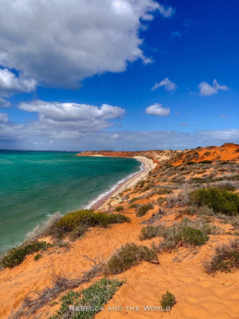 The photo captures a coastal scene at Cape Peron in Shark Bay, Western Australia. The image focuses on the sweeping curve of the coastline, where red sand dunes meet the turquoise waters of the Indian Ocean. The sky is a mix of blue and clouds, creating a picturesque view of this remote and rugged landscape.