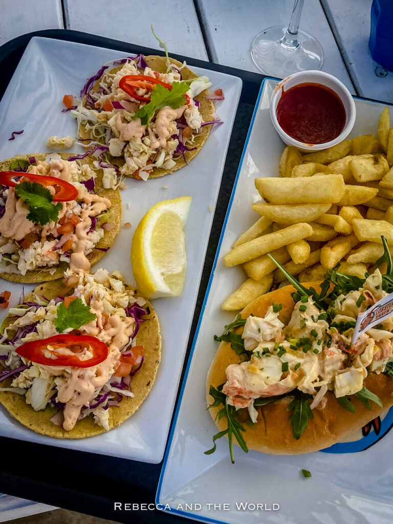 The image displays a close-up of a seafood meal served at the Lobster Shack in Cervantes, Western Australia. The meal includes lobster rolls, fries, and tacos filled with shredded lobster, topped with fresh herbs, sliced red chilli, and a drizzle of sauce. A lemon wedge and a small cup of sauce accompany the dishes, all arranged on a tray.