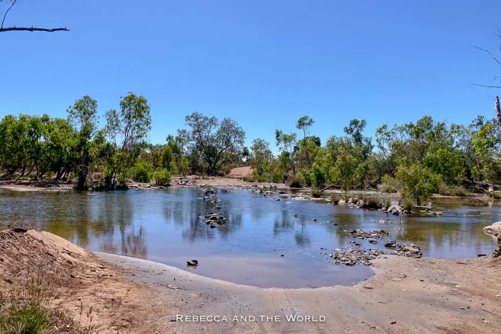 A water crossing in Purnululu National Park, where a shallow stream cuts across a dirt track surrounded by dense greenery. This image highlights the natural beauty and tranquility found within the park, as well as the need for off-road vehicles to navigate its remote areas.