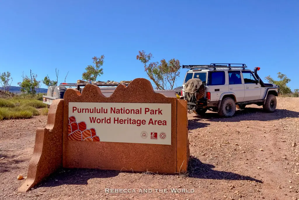 A sign at the entrance of Purnululu National Park designating it as a World Heritage Area. The sign is made of reddish stone and is accompanied by a parked off-road vehicle, indicating the rugged terrain of the park. The sign features an illustration of the Bungle Bungle Ranges.